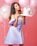 A woman in a light purple off-shoulder dress is holding a decorative cake adorned with a bow. Pink and transparent balloons with glitter are in the background, set against a pink backdrop. The cake has the name Nadia written on it.