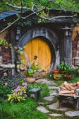 A rustic, round wooden door set into a hillside is surrounded by lush greenery and vibrant flowers. The area is adorned with potted plants and a wooden bench, while a stack of firewood is arranged on a small cart. The setting resembles a quaint, fairy-tale-like dwelling, evoking a sense of charm and mystery.