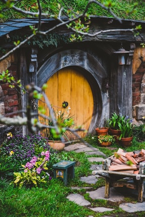 A rustic, round wooden door set into a hillside is surrounded by lush greenery and vibrant flowers. The area is adorned with potted plants and a wooden bench, while a stack of firewood is arranged on a small cart. The setting resembles a quaint, fairy-tale-like dwelling, evoking a sense of charm and mystery.