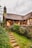 A quaint, rustic cottage with a thatched roof, surrounded by lush greenery and a small stone path leading up to its entrance. The building features wooden beams, stone chimney, and is nestled in a garden setting.