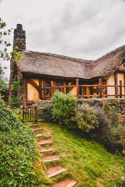 A quaint, rustic cottage with a thatched roof, surrounded by lush greenery and a small stone path leading up to its entrance. The building features wooden beams, stone chimney, and is nestled in a garden setting.