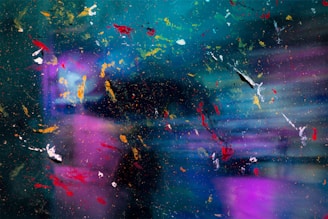 An abstract composition featuring a blend of colors and textures with splashes of vivid red, yellow, white, and orange paint scattered across a vibrant background of blue, teal, and purple tones. The image possesses a dynamic and chaotic aesthetic, resembling paint splatters or cosmic imagery.