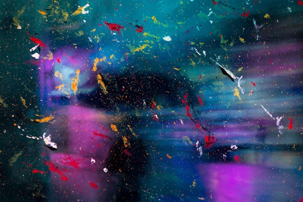 An abstract composition featuring a blend of colors and textures with splashes of vivid red, yellow, white, and orange paint scattered across a vibrant background of blue, teal, and purple tones. The image possesses a dynamic and chaotic aesthetic, resembling paint splatters or cosmic imagery.