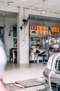 A man is sitting on a plastic chair in what appears to be an open-air food stall or hawker center. He is wearing an apron and looking at his phone. The setting features signage with Chinese and English text offering dishes like Tai Poh Mee and Satay. The background includes tiled walls, ceiling fans, and a stainless steel food counter.