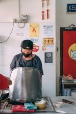 A person is preparing food behind a large metal container inside a casual dining space. The person is wearing a cap and a face mask. On the wall behind are various safety and certification signs, along with red decorations. A table in front holds a few bowls and food items.