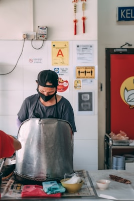 A person is preparing food behind a large metal container inside a casual dining space. The person is wearing a cap and a face mask. On the wall behind are various safety and certification signs, along with red decorations. A table in front holds a few bowls and food items.