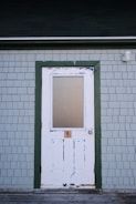 A white wooden door with peeling paint set within a green frame on a light grey shingled exterior wall. The top half of the door has a frosted glass panel. There is a lock below the glass and a round doorknob on the right side.