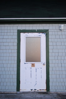 A white wooden door with peeling paint set within a green frame on a light grey shingled exterior wall. The top half of the door has a frosted glass panel. There is a lock below the glass and a round doorknob on the right side.