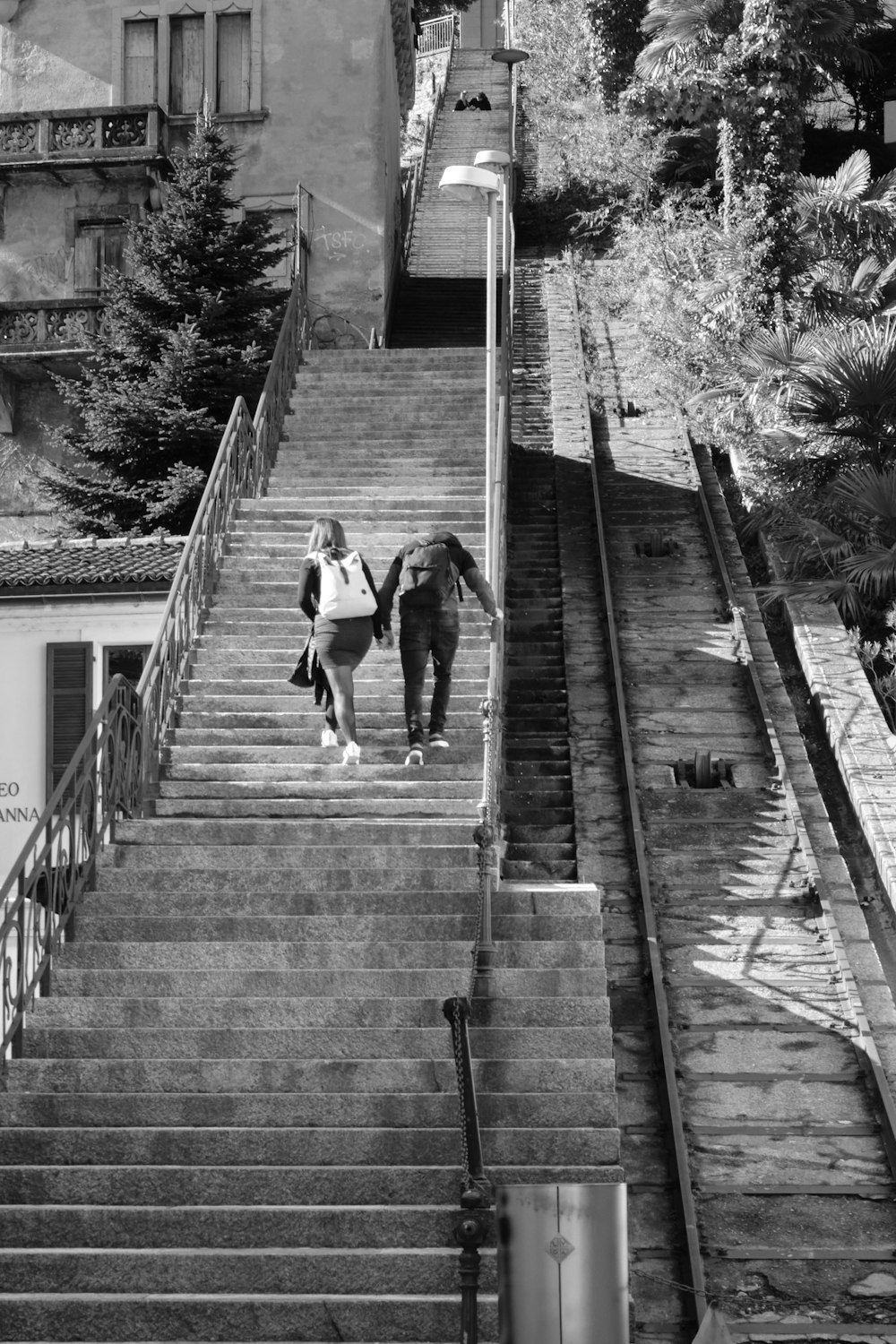 Foto en escala de grises de un hombre con camiseta negra y pantalones negros caminando sobre un puente de madera