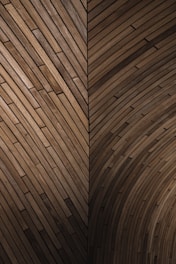 An intricate wooden pattern featuring a geometric design with overlapping planks creating a series of angled and curved sections. The wood is of a deep brown color, showcasing its natural grain and texture.