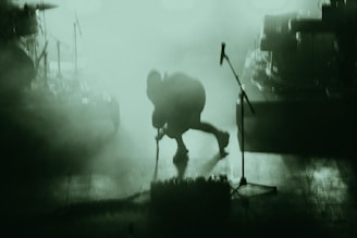 A person is on stage amidst a dense, green-tinted fog. The figure appears to be bending forward, possibly adjusting or interacting with musical equipment. Various musical instruments and equipment, such as a microphone stand and drum set, can be partially seen through the haze.
