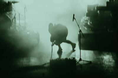 A person is on stage amidst a dense, green-tinted fog. The figure appears to be bending forward, possibly adjusting or interacting with musical equipment. Various musical instruments and equipment, such as a microphone stand and drum set, can be partially seen through the haze.