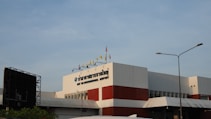 A modern airport building with a white facade featuring bold red accents. Several flags are displayed on the rooftop, including yellow and red ones. The sign on the building reads 'PHUKET INTERNATIONAL AIRPORT'. A streetlight is visible in the foreground, under a clear blue sky.