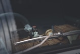 Inside a car, a small figurine is positioned on the dashboard alongside a pair of sunglasses. The dashboard mat is made of a bamboo-like material, and a folded paper napkin is visible beside the figurine. A windshield wiper is partially in view.