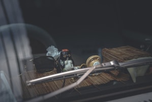 Inside a car, a small figurine is positioned on the dashboard alongside a pair of sunglasses. The dashboard mat is made of a bamboo-like material, and a folded paper napkin is visible beside the figurine. A windshield wiper is partially in view.