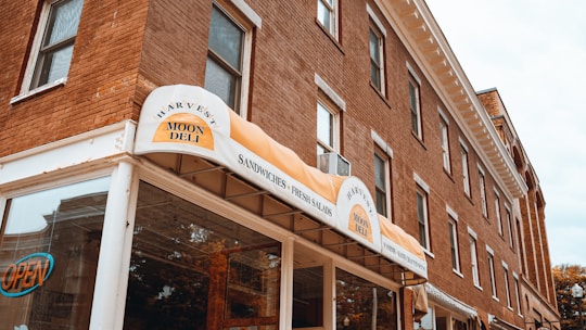 A brick building with multiple windows houses a deli named 'Harvest Moon Deli'. The entrance has an awning with signage advertising sandwiches and fresh salads. There is an 'OPEN' sign visible in one of the windows.