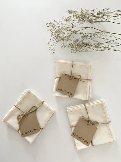 Three neatly folded stacks of beige cloth napkins tied with twine, each with a brown kraft tag labeled 'ernest & lulu.' Above them, delicate sprigs of dried flowers with small white blossoms lay against a neutral background.