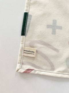 A corner of a fabric piece with an attached label displaying the text 'ernest & lulu' and a website URL. The fabric features printed abstract shapes in soft colors, bordered by stitching.