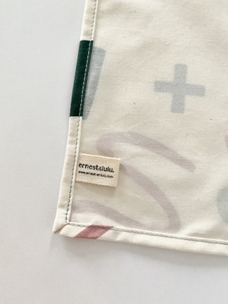 A corner of a fabric piece with an attached label displaying the text 'ernest & lulu' and a website URL. The fabric features printed abstract shapes in soft colors, bordered by stitching.