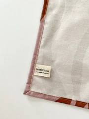 A piece of fabric is neatly laid out with visible stitching along the edges. A small label with the name 'ernestalulu' and a website URL is sewn onto the fabric near the bottom corner. The fabric has a subtle pattern and is bordered with a light brown edge.