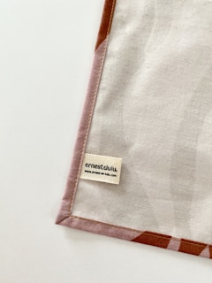 A piece of fabric is neatly laid out with visible stitching along the edges. A small label with the name 'ernestalulu' and a website URL is sewn onto the fabric near the bottom corner. The fabric has a subtle pattern and is bordered with a light brown edge.