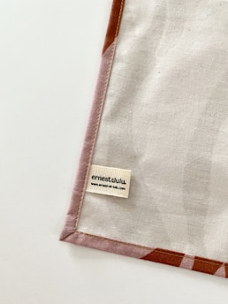 A piece of fabric is neatly laid out with visible stitching along the edges. A small label with the name 'ernestalulu' and a website URL is sewn onto the fabric near the bottom corner. The fabric has a subtle pattern and is bordered with a light brown edge.