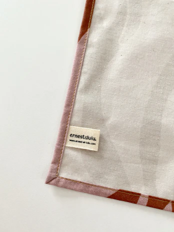 A piece of fabric is neatly laid out with visible stitching along the edges. A small label with the name 'ernestalulu' and a website URL is sewn onto the fabric near the bottom corner. The fabric has a subtle pattern and is bordered with a light brown edge.