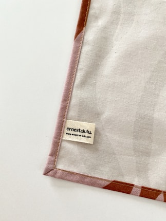 A piece of fabric is neatly laid out with visible stitching along the edges. A small label with the name 'ernestalulu' and a website URL is sewn onto the fabric near the bottom corner. The fabric has a subtle pattern and is bordered with a light brown edge.