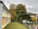 A school or office building with a yellow facade is situated beside a grassy lawn. In the background, there's a mix of green and autumn-colored trees. Overcast skies with a few patches of clouds loom above, creating a slightly overcast ambiance.