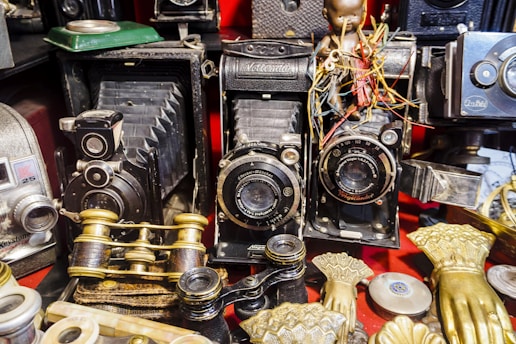 A collection of vintage cameras and binoculars is arranged among various antique items, including ornate gold-colored hand sculptures and an assortment of intricate objects. The cameras have a classical design with lenses and bellows, evoking a sense of nostalgia and historical interest.