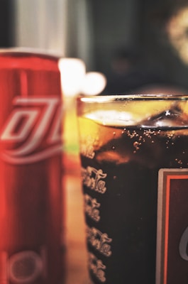 A close-up view of a glass filled with a dark, fizzy beverage with ice cubes, placed beside a red aluminum can featuring white lettering. The surroundings appear to be softly lit, creating a cozy atmosphere.