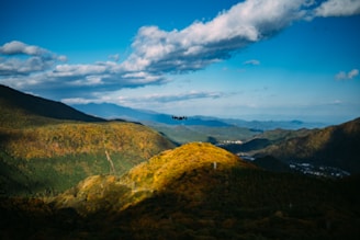 A scenic landscape featuring rolling hills covered in lush green and yellow foliage under a vibrant blue sky with scattered clouds. A black drone hovers in the center, slightly above the hills.