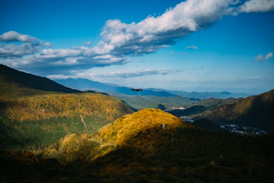 A scenic landscape featuring rolling hills covered in lush green and yellow foliage under a vibrant blue sky with scattered clouds. A black drone hovers in the center, slightly above the hills.