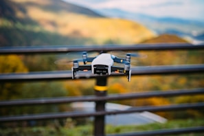 A drone is hovering outdoors against a scenic backdrop of hills and a blurred background. The focus is on the drone, with its rotors spinning and a metallic finish.