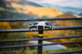 A drone is hovering outdoors against a scenic backdrop of hills and a blurred background. The focus is on the drone, with its rotors spinning and a metallic finish.