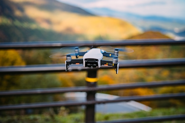 A drone is hovering outdoors against a scenic backdrop of hills and a blurred background. The focus is on the drone, with its rotors spinning and a metallic finish.