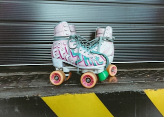 A pair of retro-style roller skates featuring colorful designs is positioned against a textured metal backdrop. The skates have a graffiti-like pattern with bold lettering on the sides, and each skate sits on four wheels. The background includes horizontal lines and a safety warning pattern with black and yellow stripes at the bottom.