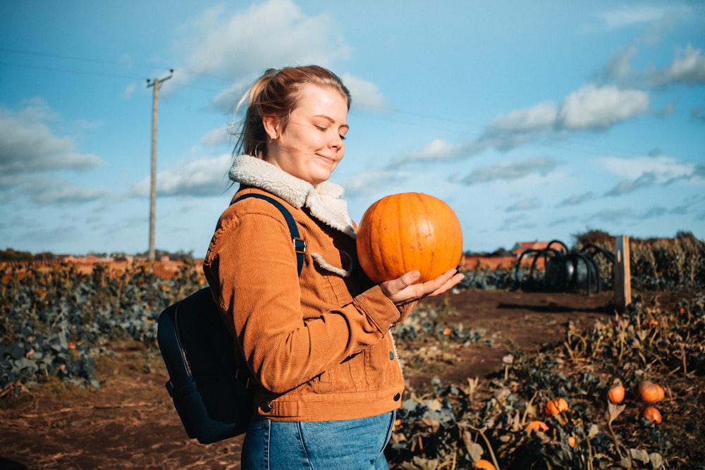 girl in brown jacket and blue denim jeans holding pumpkin during daytime