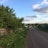 A rural road stretches into the distance, bordered by green grass and lush trees on one side and a wooden fence on the other. Small houses with colorful roofs are visible along the road, under a sky partially covered with fluffy clouds.