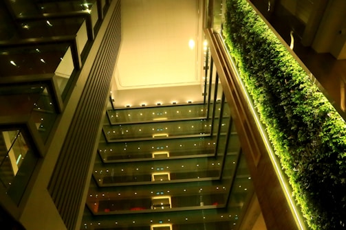 An interior view of a modern building with multiple levels. The design features a vertical garden with lush greenery on one side. The opposite wall contains glass and metal elements, with visible lights that illuminate the structure. The overall architecture is sleek and contemporary.