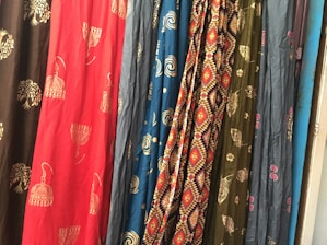 Several colorful fabrics are hanging side by side, each with unique patterns including geometric designs, floral motifs, and abstract shapes. The textures and folds of the materials create a rich visual tapestry.