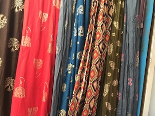 Several colorful fabrics are hanging side by side, each with unique patterns including geometric designs, floral motifs, and abstract shapes. The textures and folds of the materials create a rich visual tapestry.
