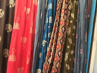 Several colorful fabrics are hanging side by side, each with unique patterns including geometric designs, floral motifs, and abstract shapes. The textures and folds of the materials create a rich visual tapestry.