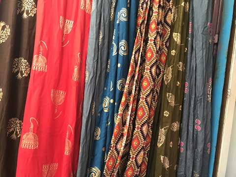 Several colorful fabrics are hanging side by side, each with unique patterns including geometric designs, floral motifs, and abstract shapes. The textures and folds of the materials create a rich visual tapestry.
