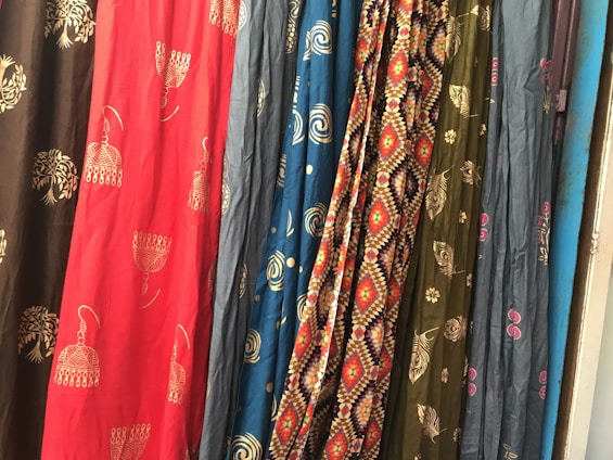 Several colorful fabrics are hanging side by side, each with unique patterns including geometric designs, floral motifs, and abstract shapes. The textures and folds of the materials create a rich visual tapestry.