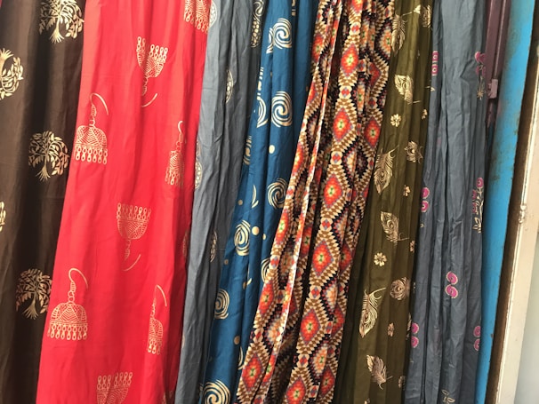 Several colorful fabrics are hanging side by side, each with unique patterns including geometric designs, floral motifs, and abstract shapes. The textures and folds of the materials create a rich visual tapestry.