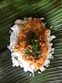 A serving of rice topped with a thick, spicy orange-colored sauce containing chopped herbs and small crispy pieces, placed on a large green leaf, likely a banana leaf, which serves as a natural plate.