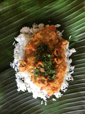 A serving of rice topped with a thick, spicy orange-colored sauce containing chopped herbs and small crispy pieces, placed on a large green leaf, likely a banana leaf, which serves as a natural plate.