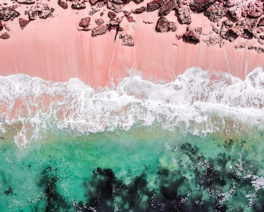 Aerial view of a coastline with pink sand transitioning into foamy white waves meeting the turquoise ocean. Large rocks are scattered along the pink beach, creating a stark contrast with the smooth sand and vibrant water.