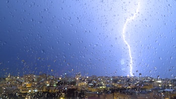 A vibrant lightning bolt pierces the night sky, illuminating a cityscape in the background. Raindrops scatter across a windowpane, capturing the dramatic weather outside and showcasing the city lights.